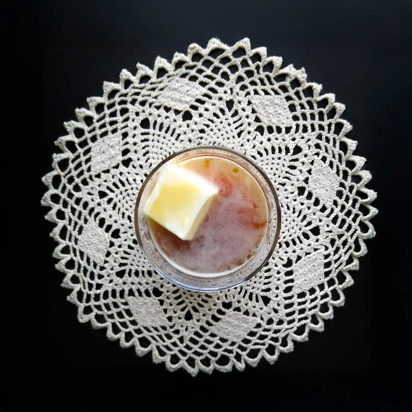 Overhead shot of a pinkish-red cocktail topped with a pat of butter in a rocks glass set on a white crocheted doily