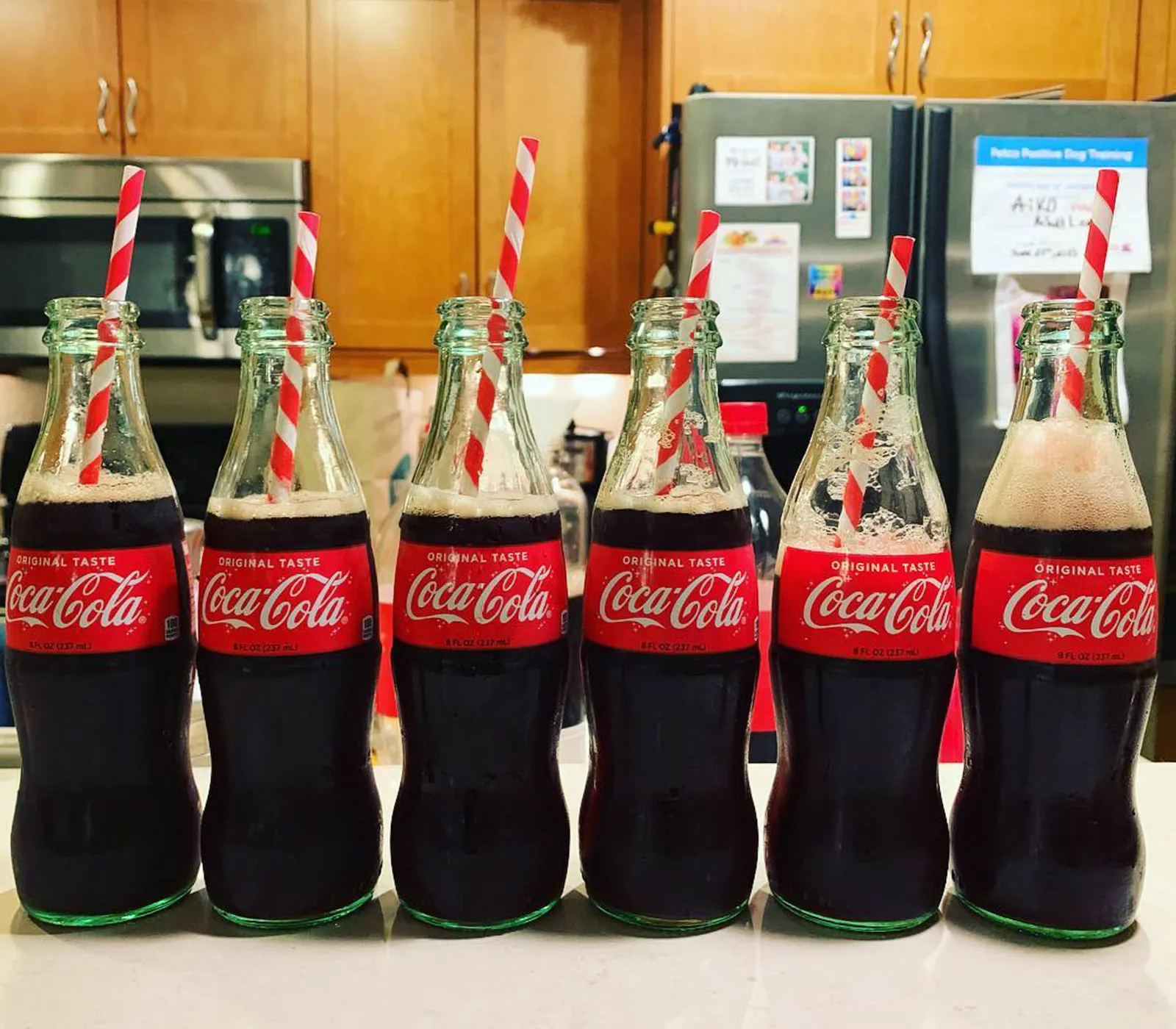 Six glass Coca-Cola bottles fitted with red and white striped straws lined up on a kitchen counter