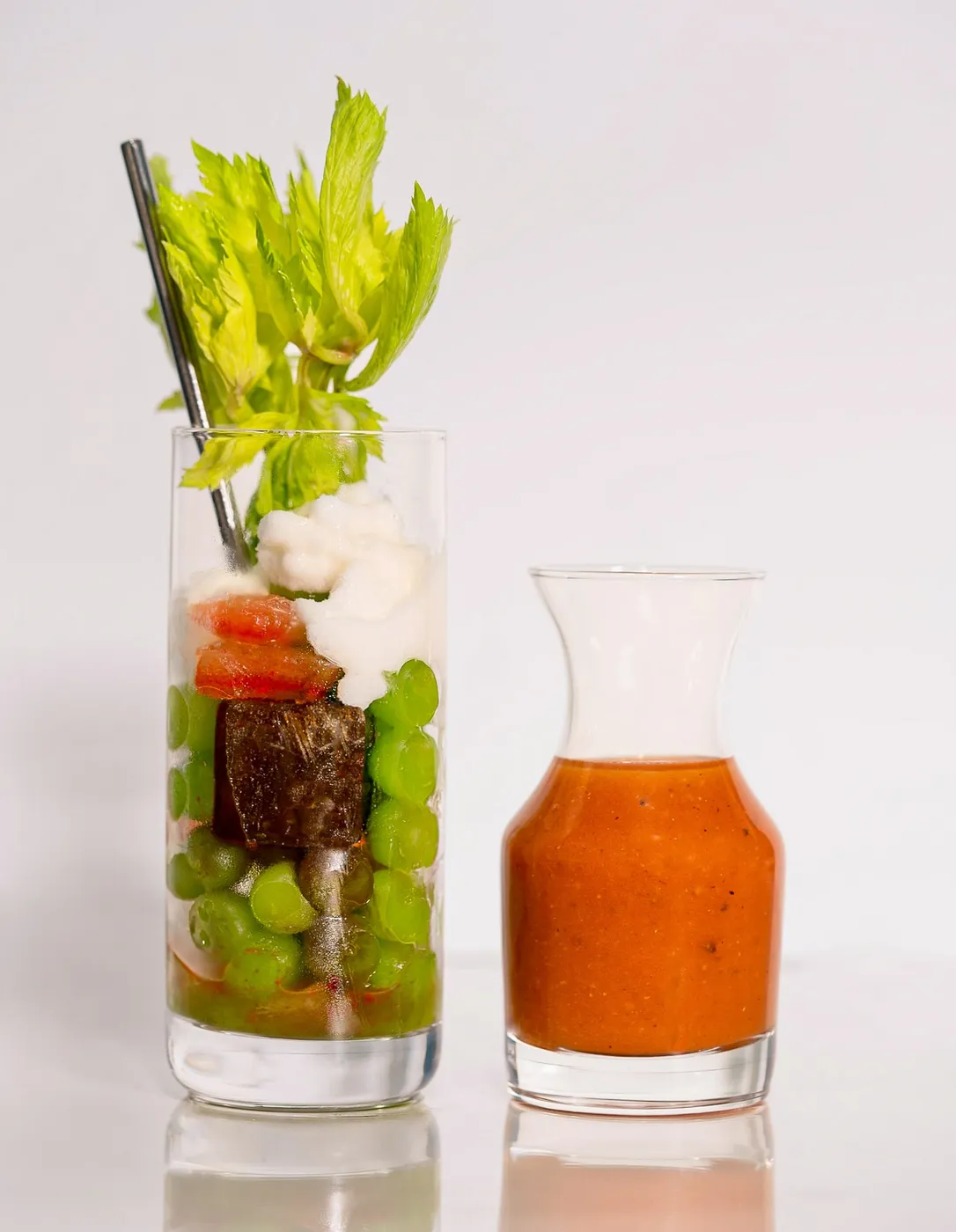 A Bloody Mary layered with green grapes, seared beef, tomato, and foam, garnished with celery, beside a small carafe of Bloody Mary mix