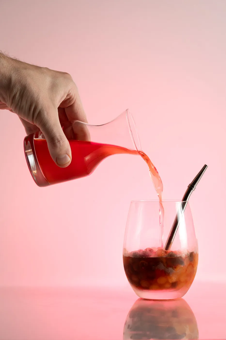 A hand pouring a pink cocktail from a red-tinted decanter into a stemless glass filled with berries and ice, against a pink background