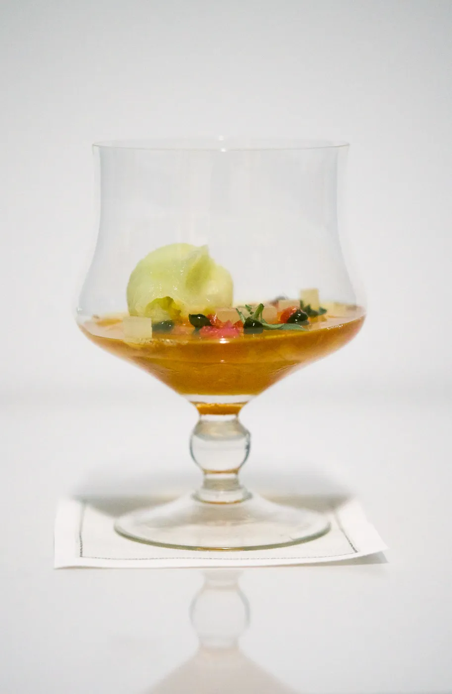 An amber cocktail in a coupe topped with a pale green sorbet quenelle and scattered edible garnishes