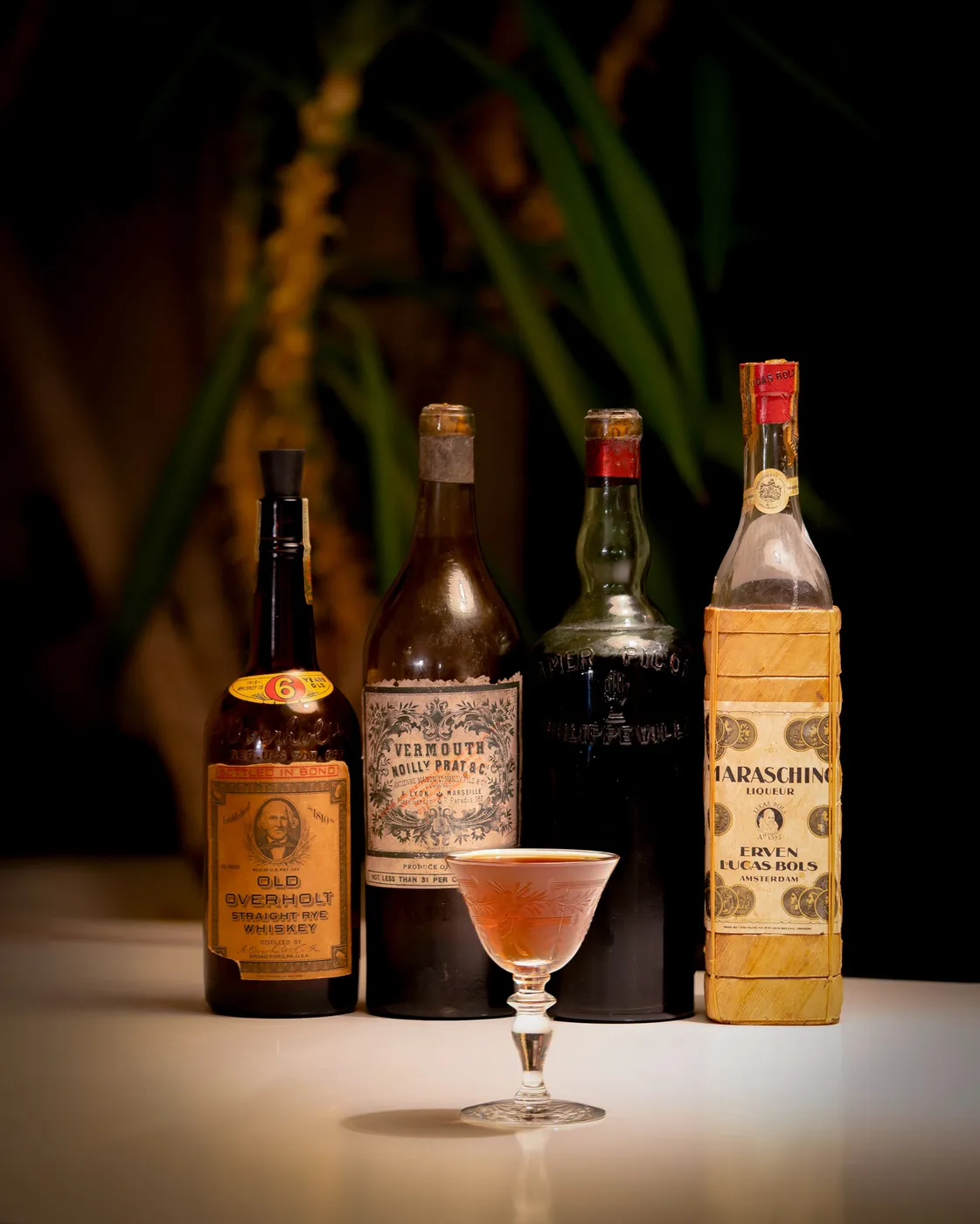 A Brooklyn cocktail in an etched coupe glass in front of vintage bottles of Old Overholt rye, Noilly Prat vermouth, Amer Picon, and Bols maraschino liqueur
