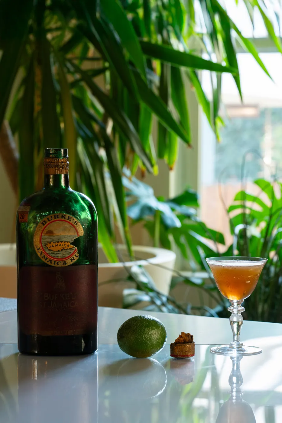 A vintage bottle of Burke's Jamaica Rum beside a lime and a coupe glass of amber daiquiri, surrounded by houseplants