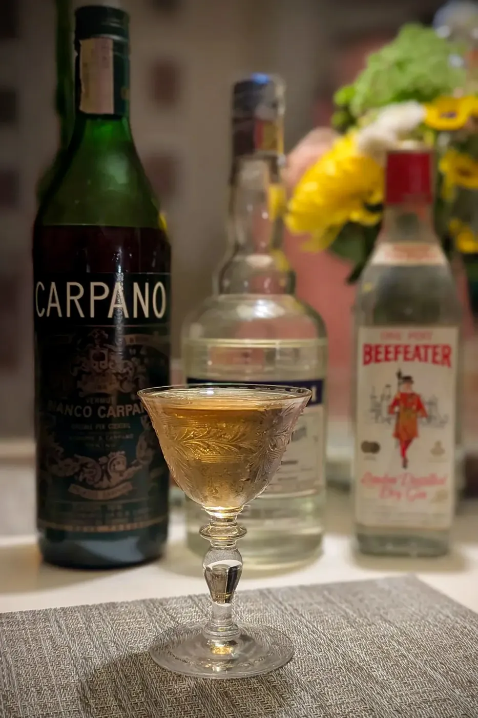 A pale gold cocktail in a coupe glass in front of vintage bottles of Carpano vermouth and Beefeater gin, with yellow flowers blurred in the background