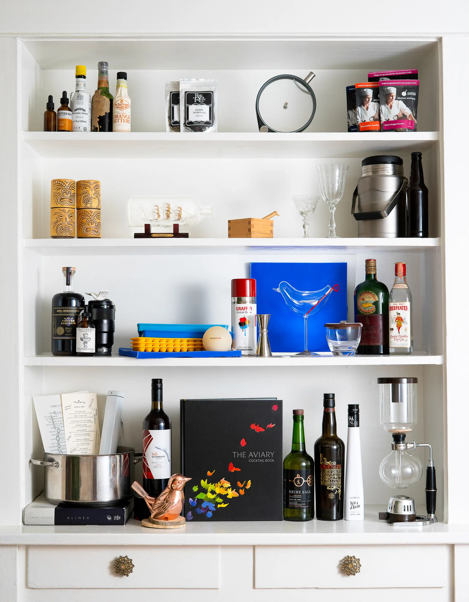 A white built-in cabinet stocked with bar tools, tiki mugs, bitters, a ship in a bottle, a blue glass bird, and a copy of The Aviary Cocktail Book