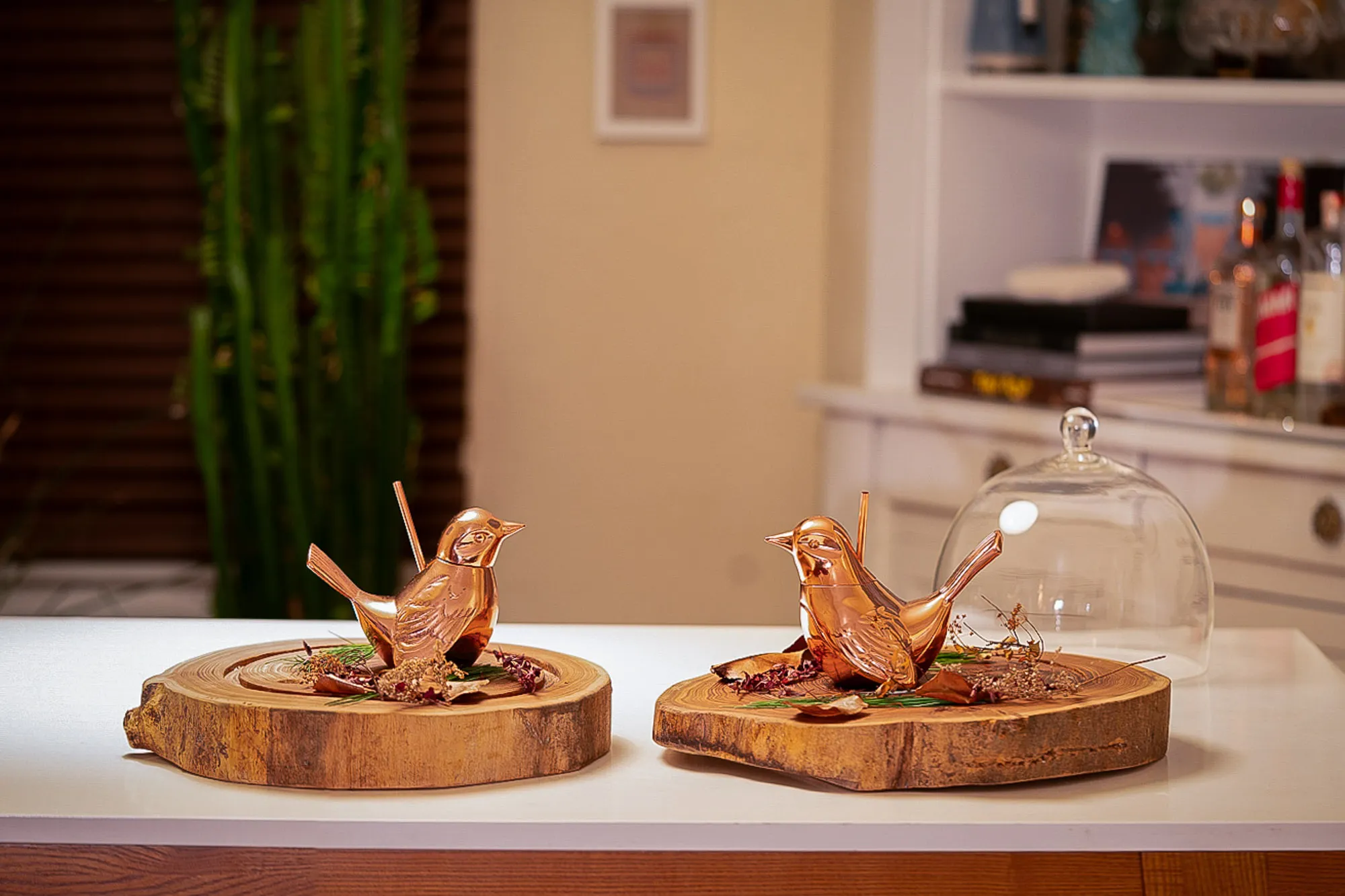 Two copper bird-shaped cocktail vessels with straws, perched on wood slices with dried flowers, one covered by a glass dome