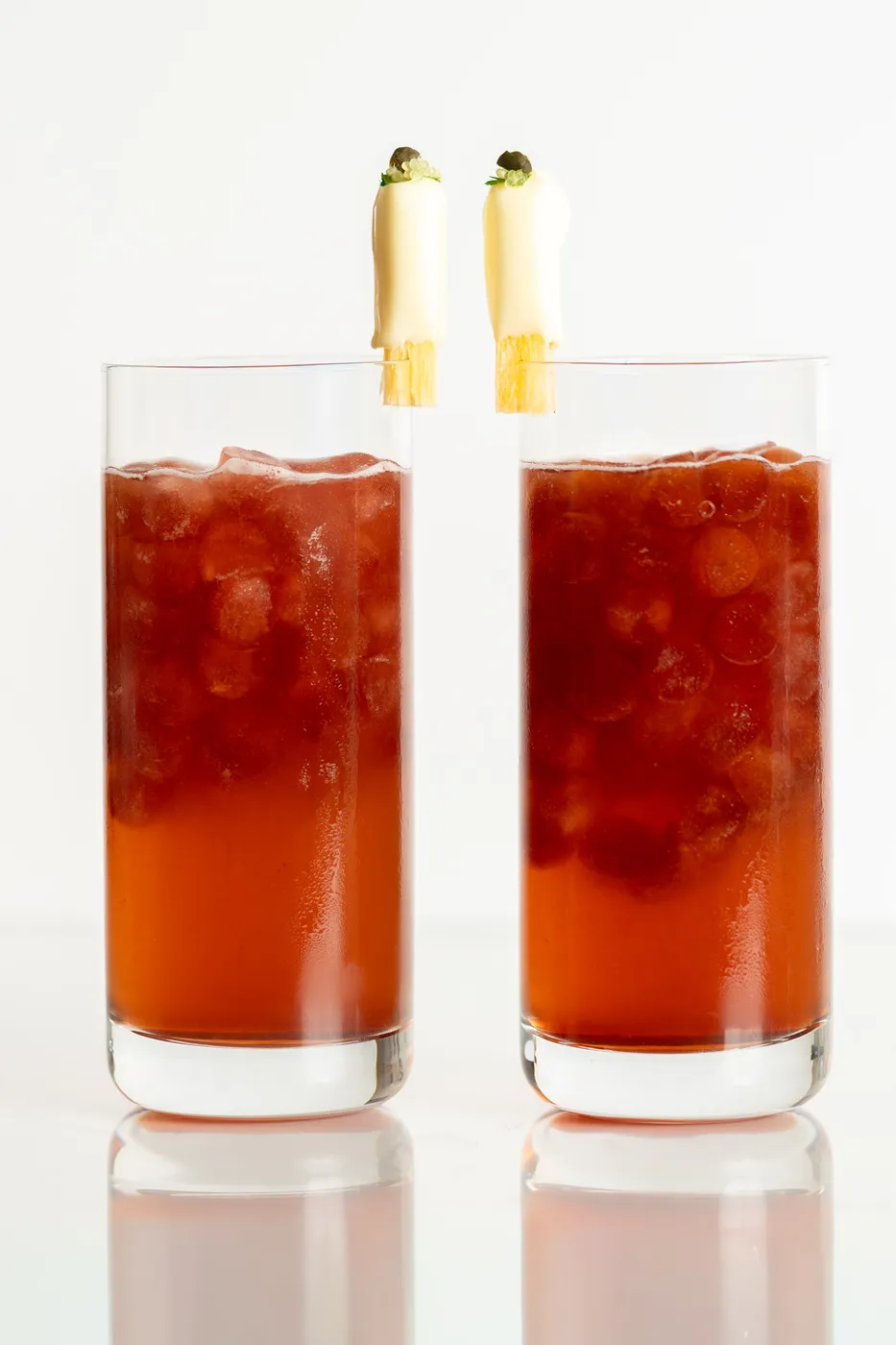 A pair of red carbonated cocktails in highball glasses garnished with white chocolate sugar cane segments