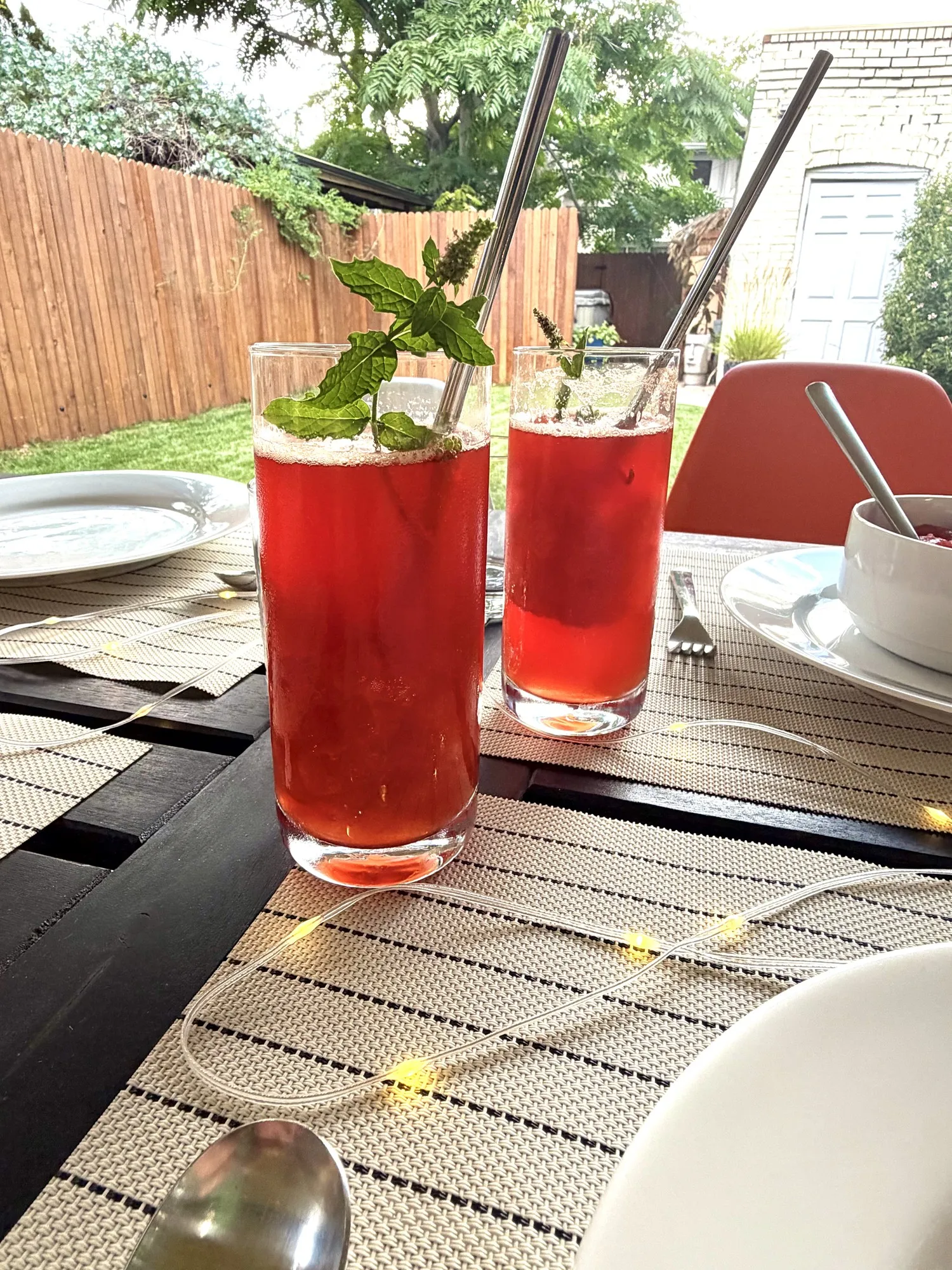 Two tall glasses of red cocktails garnished with mint and metal straws on an outdoor patio table, surrounded by rope lights.