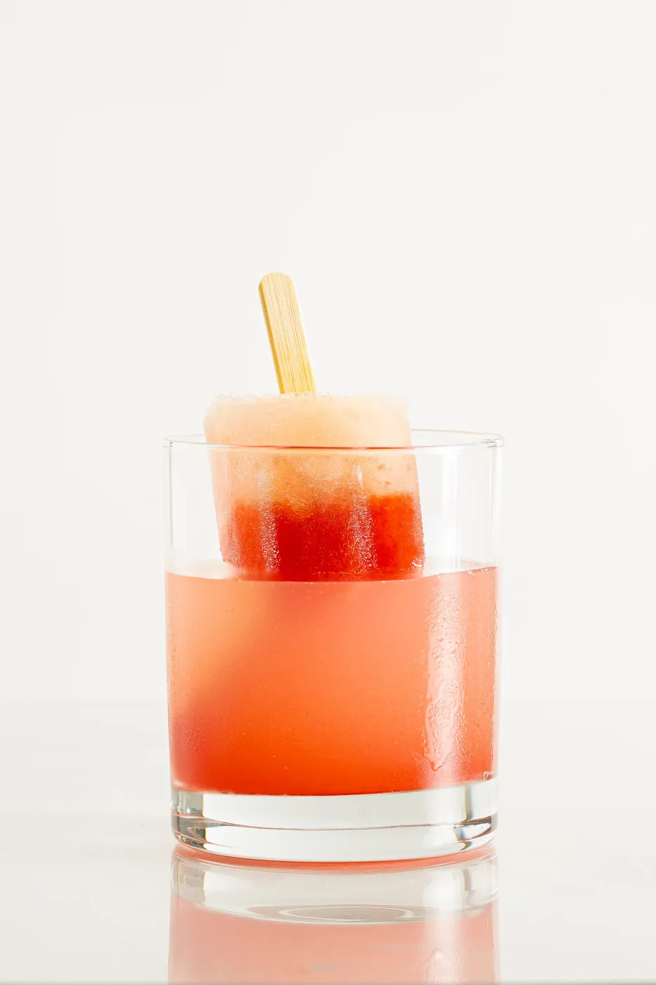 A rocks glass of orange-red cocktail garnished with a half-melted watermelon-colored popsicle, against a white background