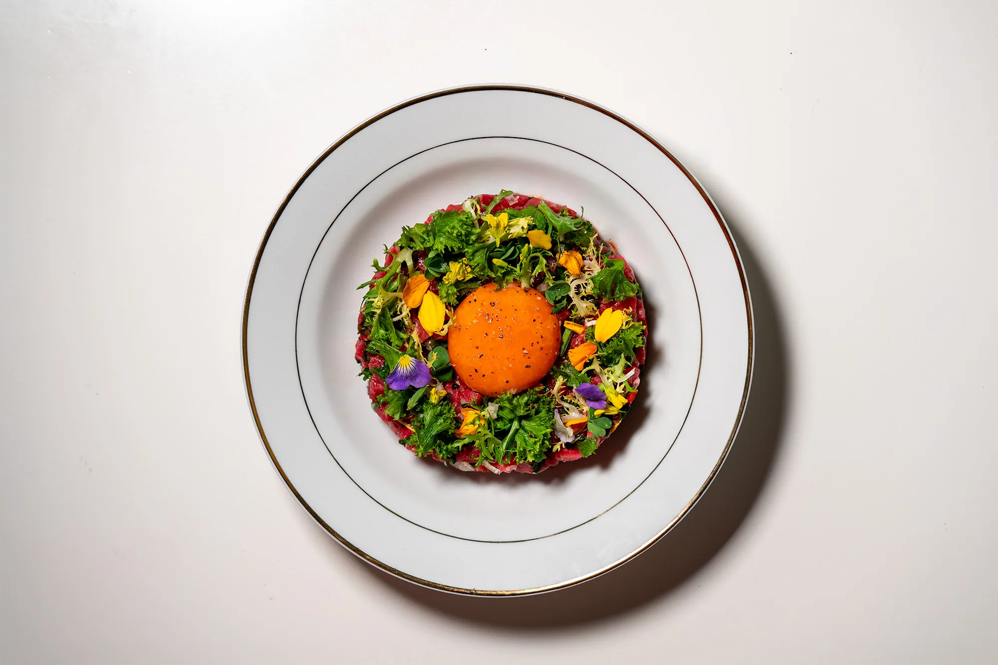 An overhead view of beef tartare topped with a bright orange egg yolk and edible flowers