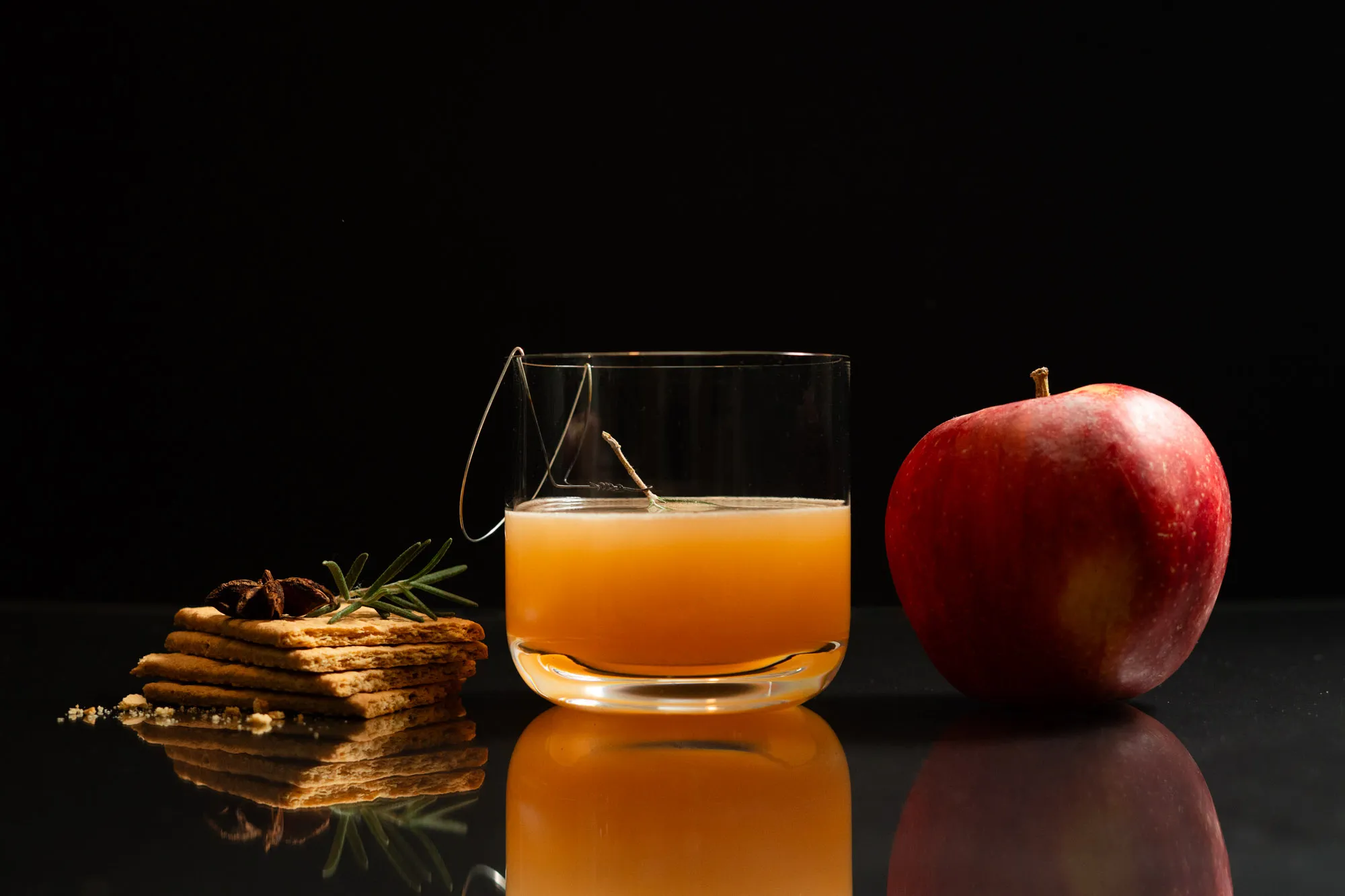 An amber cocktail garnished with a rosemary sprig and star anise atop crackers on a wire, beside a red apple