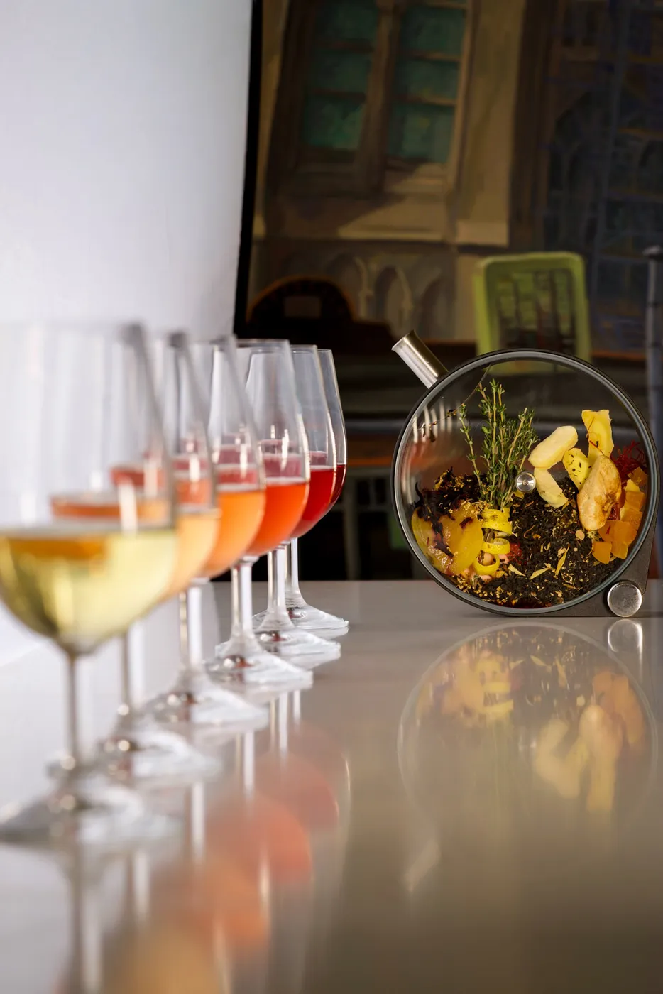 A row of cocktails in graduating amber and red shades beside a round glass tea-infusion chamber packed with loose tea leaves and dried fruit