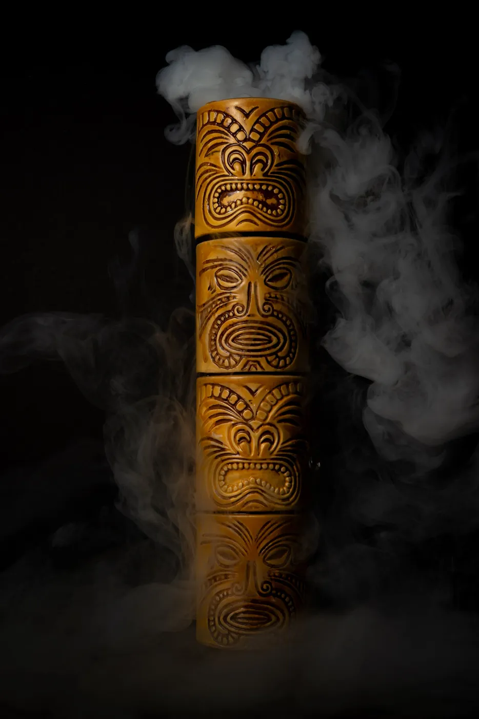 A carved wooden tiki mug with three totem faces, wreathed in rising smoke