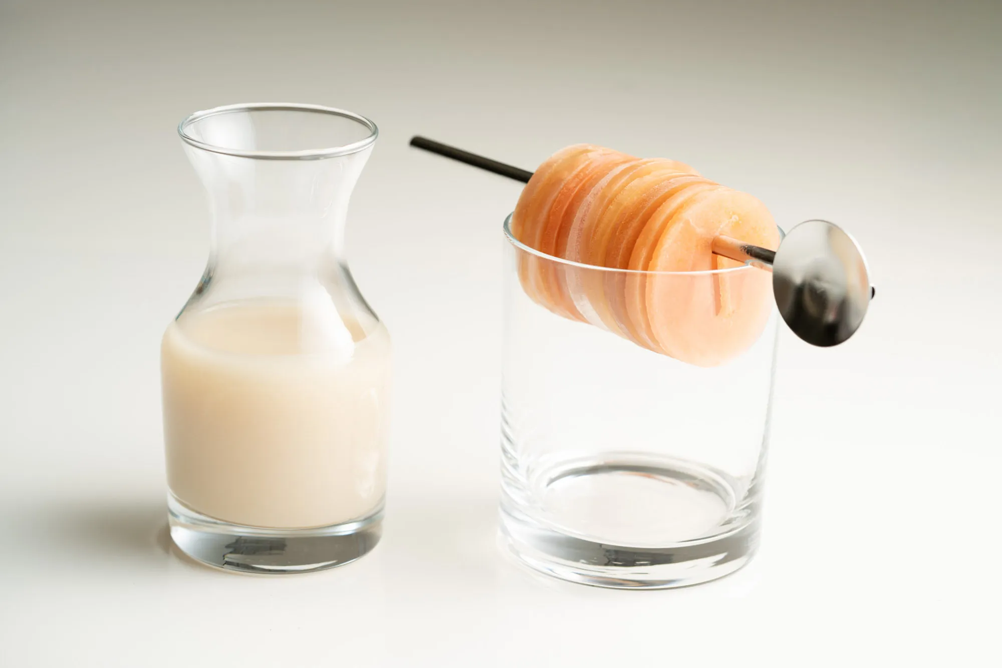 A carafe of pale creamy liquid beside a rocks glass holding a stack of translucent orange ice discs threaded on a bar spoon
