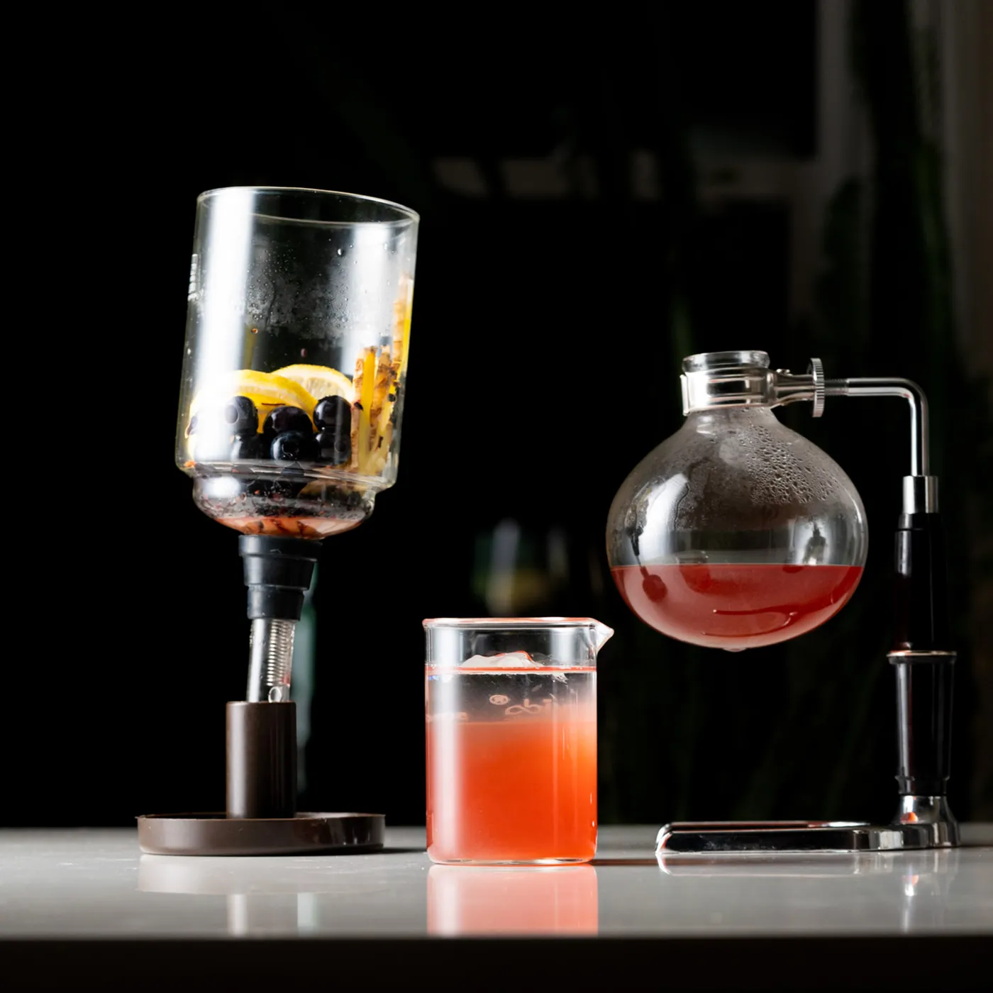 A vacuum pot cocktail setup with citrus, blueberries, and botanicals in the upper chamber and red-orange liquid extracting into the lower globe, beside a beaker of the finished drink
