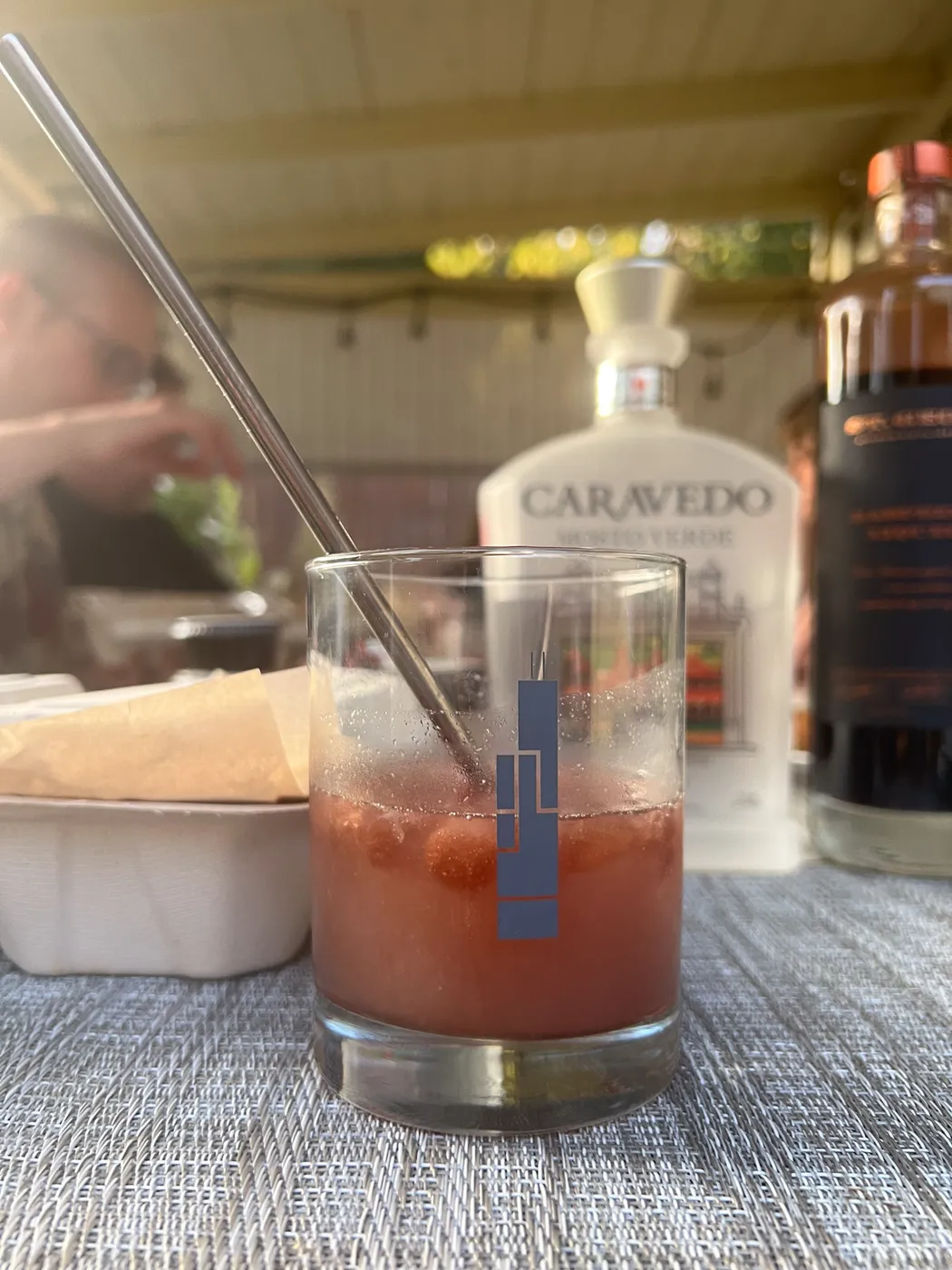 A reddish cocktail with a metal straw in a rocks glass on an outdoor table, with liquor bottles blurred in the background